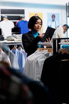 Clothing store asian woman employee using digital tablet while working in shopping center. Department mall boutique worker examining apparel in stock and managing inventory