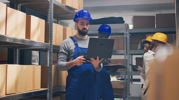 Caucasian man preparing delivery order with laptop, examining cardboard boxes with merchandise and stock. Warehouse manager in overalls and hardhat working in storage room, small business.