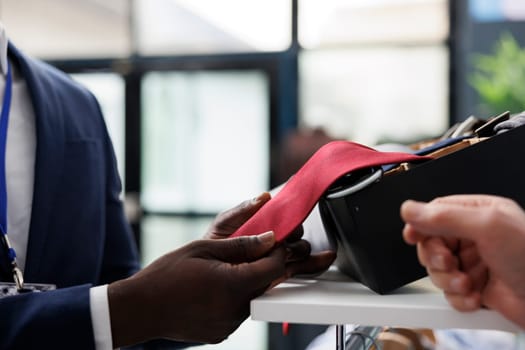 Stylish employee helping elderly shopper with red tie, checking fabric in shopping centre. Senior client buying trendy clothes and fashionable accessories for casual wear in modern boutique. Close up