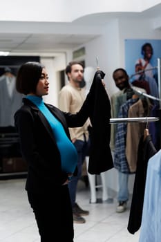 Pregnant asian customer examining clothing rack and holding jacket on hanger in department store. Future mother holding belly and choosing maternity apparel in fashion boutique