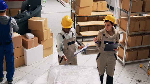 Depot workers planning shipment and distribution in warehouse space, analyzing list of stock inventory. Storage room manager organizing industrial goods in cardboard boxes, small business.