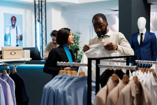 Clothing store african american customer holding shirt on hanger, discussing style and fit with consultant. Shopping mall showroom asian woman assistant offering client advice