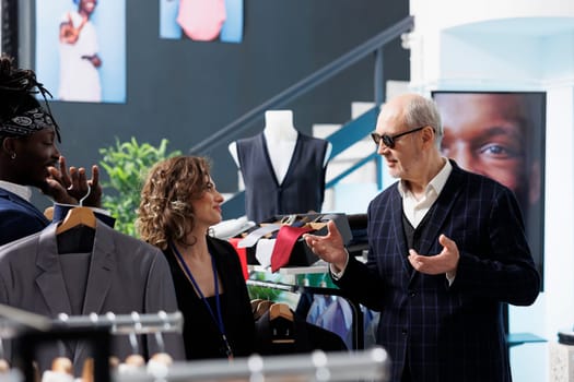 Showroom employees helping elderly customer with trendy shirt, choosing stylish clothes for formal wear in shopping centre. Senior man buying fashionable merchandise and accessories in modern boutique