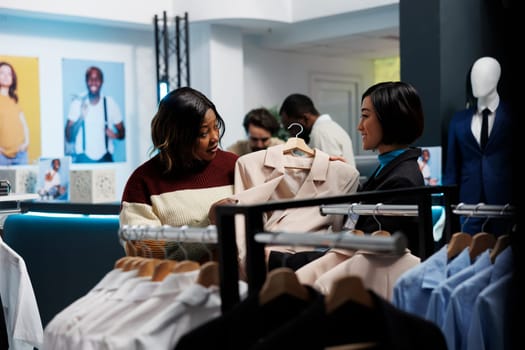 African american woman examining jacket and asking clothing store assistant for advice. Mall fashion boutique customer talking with consultant while shopping for trendy outfit