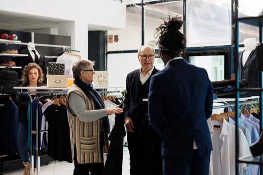 Elderly couple looking to buy formal wear, asking store manager about clothes fabric in modern boutique. Senior man buying fashionable merchandise and stylish accessories in shopping centre