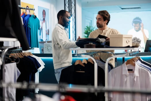 Young caucasian man searching for casual menswear and chatting with shopping center employee. Customer selecting casual apparel and accessory and asking store worker for recommendation