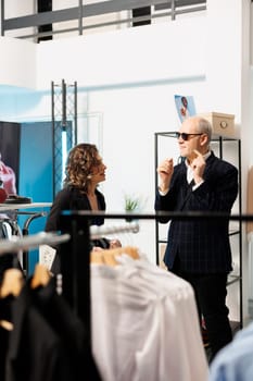 Senior man trying stylish sunglasses in clothing store, needed help from showroom employee. Elderly customer looking at new fashion collection, buying fashionable clothes in modern boutique