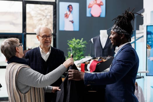 Old couple checking shirt material with boutique employee, buying trendy clothes for casual wear in clothing centre. Elderly customer looking at fashion collection in shopping centre. Fashion concept