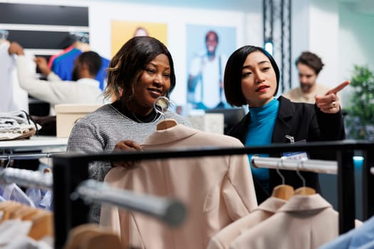 Clothing store asian employee pointing with finger and showcasing apparel options to buyer. Smiling african american woman holding jacket on hanger while shopping in department mall