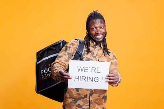 Takeaway delivery worker with thermal backpack holding job hire sign for courier position. African american man wanting perfect candidate for career opportunity in studio over yellow background