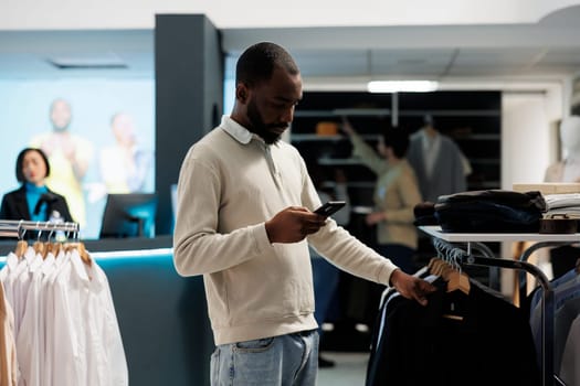 African american man scrolling clothing store online website on smartphone while shopping in department mall. Boutique customer checking shirt size in stock using mobile phone app