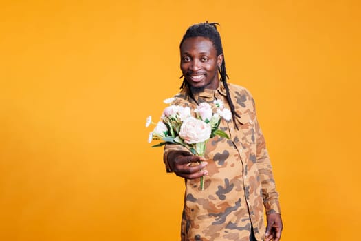 Portrait of african american man holding white flowers bouquet, presenting surprise for girlfriend at camera in studio over yellow background. Romantic boyfriend posing for valentine's day