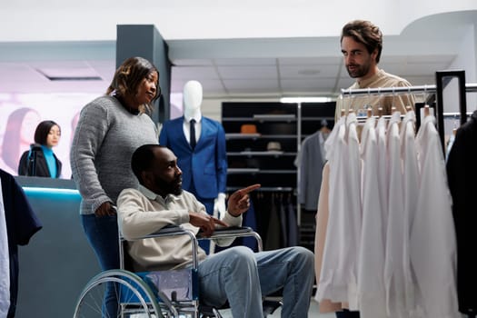 African american man with chronic impairment receiving advice from clothing store assistant while shopping with friend. Department mall customer in wheelchair talking with consultant