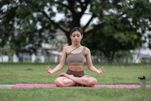 Young Asian woman doing yoga, meditation on a calm yoga mat in a green park in the early morning. Young woman wearing brown yoga clothes Yoga concept.