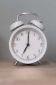 Close-up of a round white alarm clock on a table in the bedroom. Blurred photo. The hands on the clock show seven o'clock in the morning, time to get up. Retro alarm clock on the table, vintage tone.