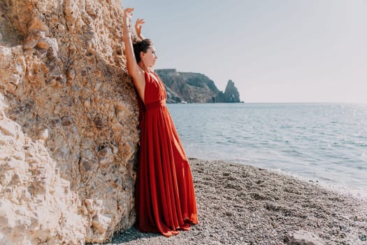 Side view a Young beautiful sensual woman in a red long dress posing on a rock high above the sea during sunrise. Girl on the nature on blue sky background. Fashion photo.