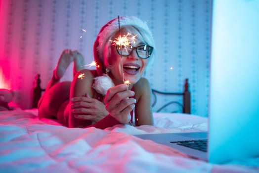 Sexy girl in lingerie wearing a Santa Claus hat poses sexily on a bed with a laptop by a Bengal fire on Christmas Day in neon lights
