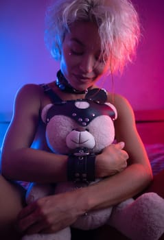 sexy girl in leather in a bdsm accessory on the bed with a cute teddy bear with emotions and in neon light