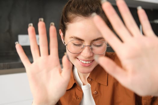 Portrait of cute and romantic young woman in glasses, covers her face, extends hands to block camera, asks to stop taking pictures.