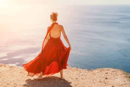 Side view a Young beautiful sensual woman in a red long dress posing on a rock high above the sea during sunrise. Girl on the nature on blue sky background. Fashion photo.