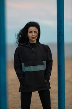 A beautiful brunette fitness model is working out on a sports ground by the sea. Young woman posing in an anorak and leggings outdoors. Fashionable sportswear for street training