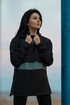 A beautiful brunette fitness model is working out on a sports ground by the sea. Young woman posing in an anorak and leggings outdoors. Fashionable sportswear for street training