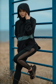 A beautiful brunette fitness model is working out on a sports ground by the sea. Young woman posing in an anorak and leggings outdoors. Fashionable sportswear for street training