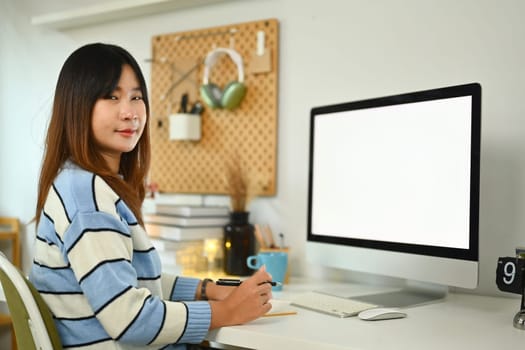 Portrait of young Asian woman sitting in front of computer monitor and smiling at camera.