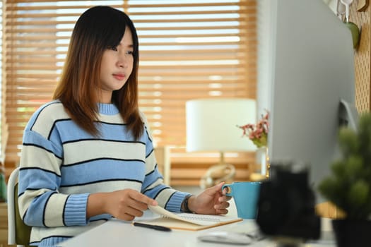 Concentrate young woman in sweater looking at computer monitor while working online at home.