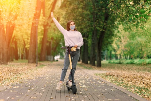 Casual caucasian female wearing protective face mask riding urban electric scooter in city park during covid pandemic. Urban mobility concept.