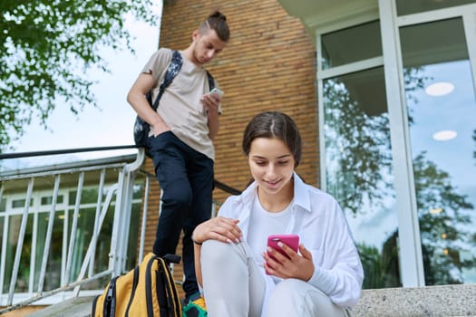 Teenage students with smartphones on the steps of educational building, in focus teenager girl high school age using gadget. Adolescence, youth, education, lifestyle, technology concept