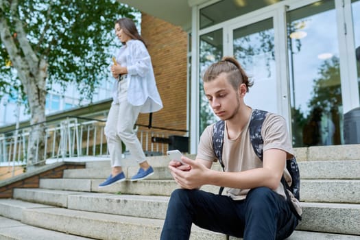 Hipster guy teenager student 18, 19 years old with backpack using smartphone, sitting on steps of outdoor educational building. Youth, education, lifestyle, technology