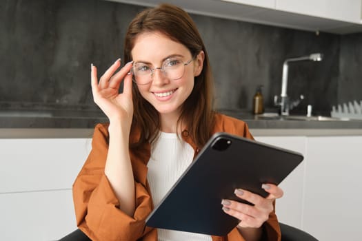 Portrait of beautiful young female model in glasses, holding digital tablet, wearing glasses, reading e-book, posing in kitchen at home.