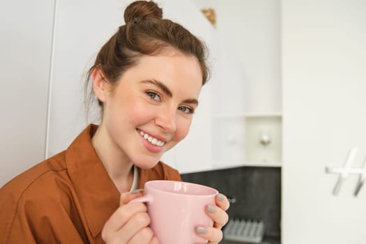 Close up portrait of beautiful brunette woman with cup of coffee, drinking aromatic beverage from mug, smiling from pleasure, sitting in kitchen at home.