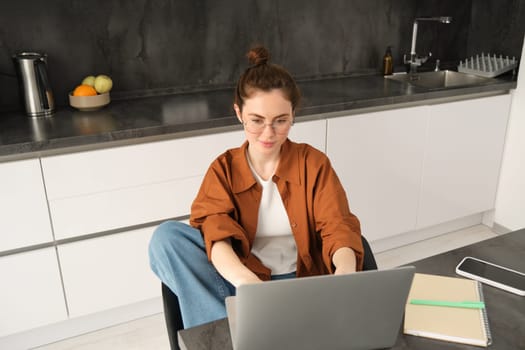 Portrait of beautiful young woman studying at home, sitting with laptop and documents in kitchen. Self-employed business owner typing on computer, sending emails to clients.