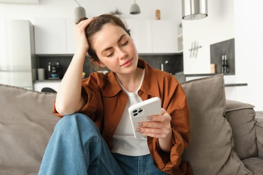 Portrait of beautiful young woman reading news on mobile phone, using smartphone app, looking at screen and smiling, sitting at home on sofa.