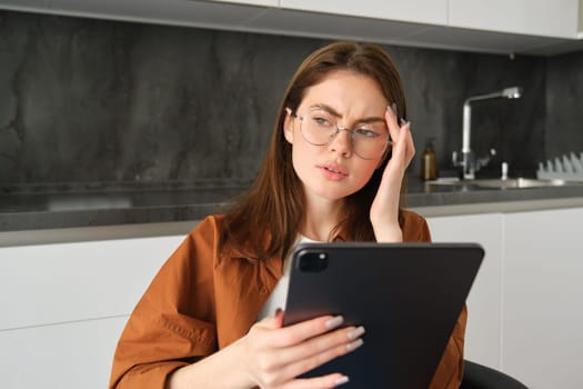 Portrait of woman freelancer, wearing glasses, holding digital tablet, looking tired, has migraine or headache, reading e-book. Freelance worker sits at home with gadget.