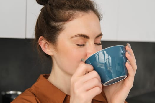 Portrait of beautiful young woman, enjoying delicious aroma of freshly made coffee, holding mug, standing near machine in the kitchen, drinking from mug.