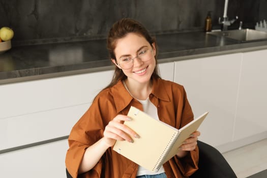 Close up portrait of young woman, student in kitchen, holding notebook, revising for exam at home, studying, reading her planner.