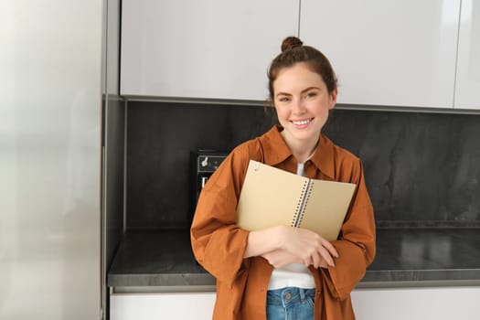 Portrait of beautiful modern woman, standing in the kitchen, flipping recipe book, holding notebook and smiling, reading notes.