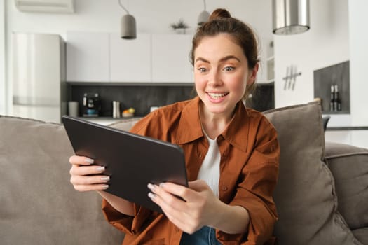 Portrait of excited, smiling young woman playing games on her digital tablet, tilting gadget and looking amused at screen, sitting on couch at home.