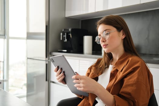 Home and leisure concept. Portrait of beautiful young woman reading on digital tablet, sitting in kitchen, working on remote.