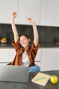 Portrait of cheerful young woman, student stretches her hands with pleased face, accomplish goal, finish working on project, using laptop and smiling pleased.