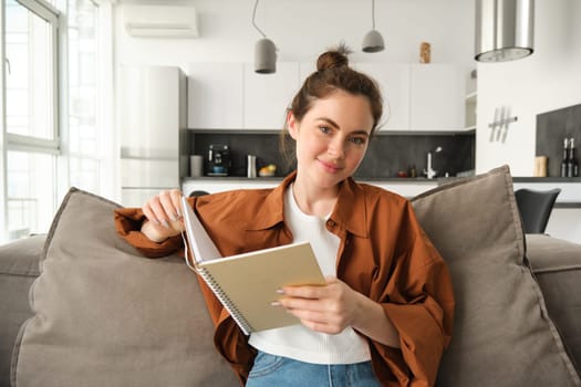 Beautiful young female student, sitting at home on sofa, relaxing on couch, reading notebook and smiling.