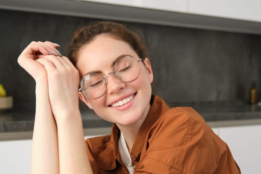 Close up portrait of brunette woman in glasses, beautiful female model sitting in kitchen, laughing and smiling, express carefree, positive emotion.