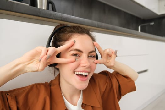 Close up of carefree young woman, laughing and smiling, showing peace, v-sign gesture, listening to music in wireless headphones.