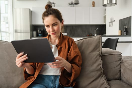 Portrait of smiling beautiful woman, sitting on couch, watching videos on digital tablet, reading e-book on her device, resting at home.