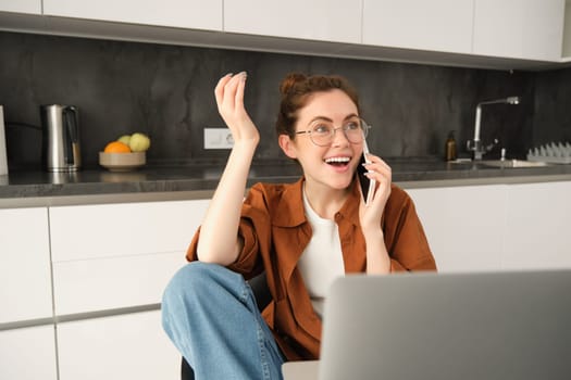 Portrait of young professional, business owner at home, talking on mobile phone, calling coworker while working on remote. Student studying and answer telephone, sitting near laptop.
