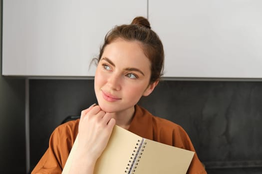 Portrait of young modern woman standing in the kitchen with notebook, reading documents, smiling and looking happy.