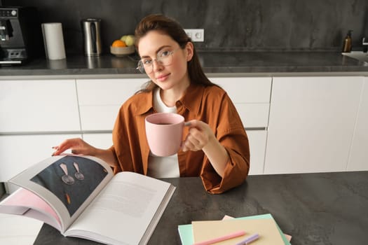 Portrait of young woman enjoying her weekend, reading a book and drinking tea at home, wearing glasses and casual clothes.